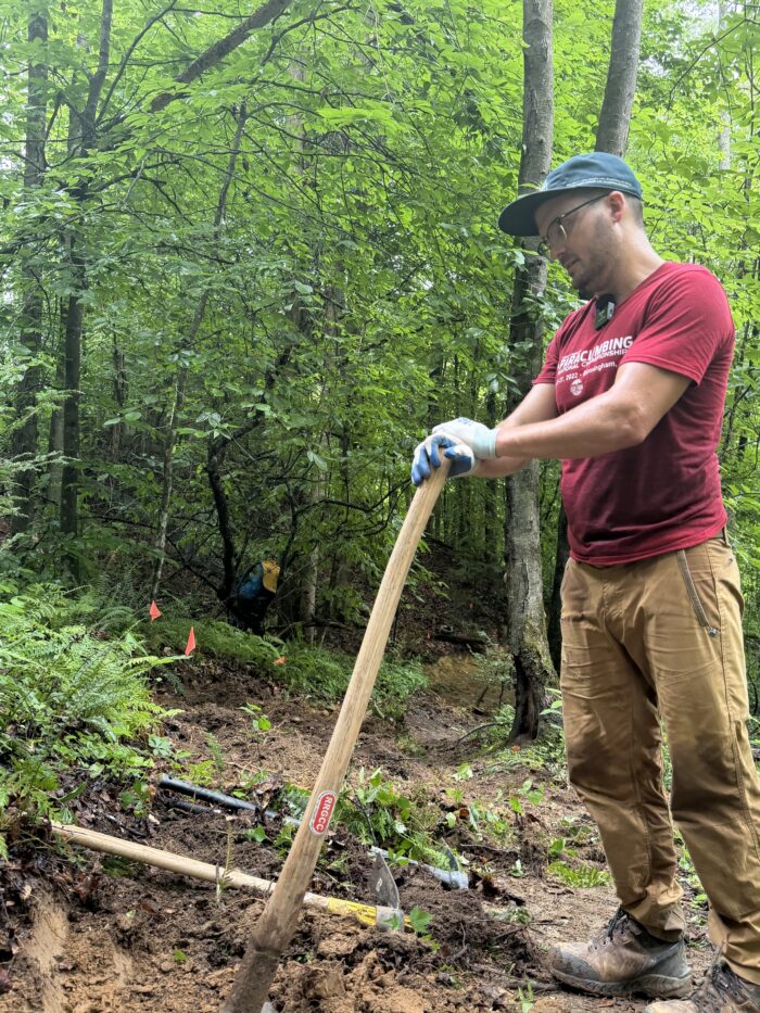 A man wearing a ball cap, red shirt, tan pants, glasses, and gloves rests his hands on a pickaxe.