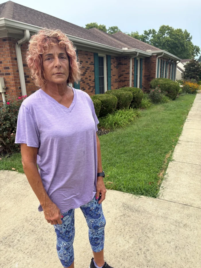 A woman stands in front of a house. She is not smiling. She wears blue leggings and a light purple shirt.