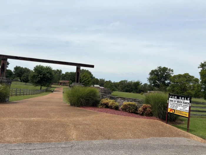 A driveway into a beautiful looking home. A for sale sign is seen at the entrance.