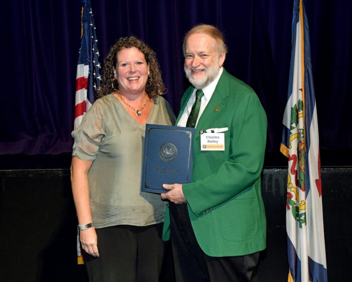 Man in green jacket holds up a blue folder with WV state seal on it.
