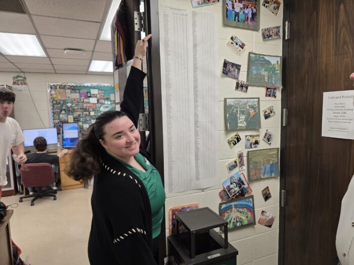 young woman in green and black points at her name on a long list on the wall