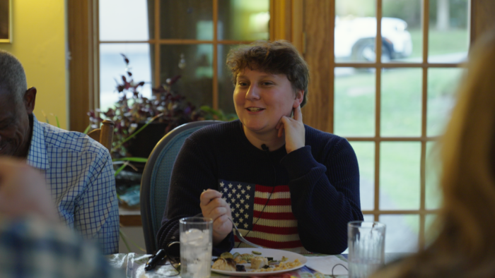 A younger woman with brown, pixie cut hair. She wears a sweater featuring the American flag prominently on the front. She is looking off camera and appears to be speaking.