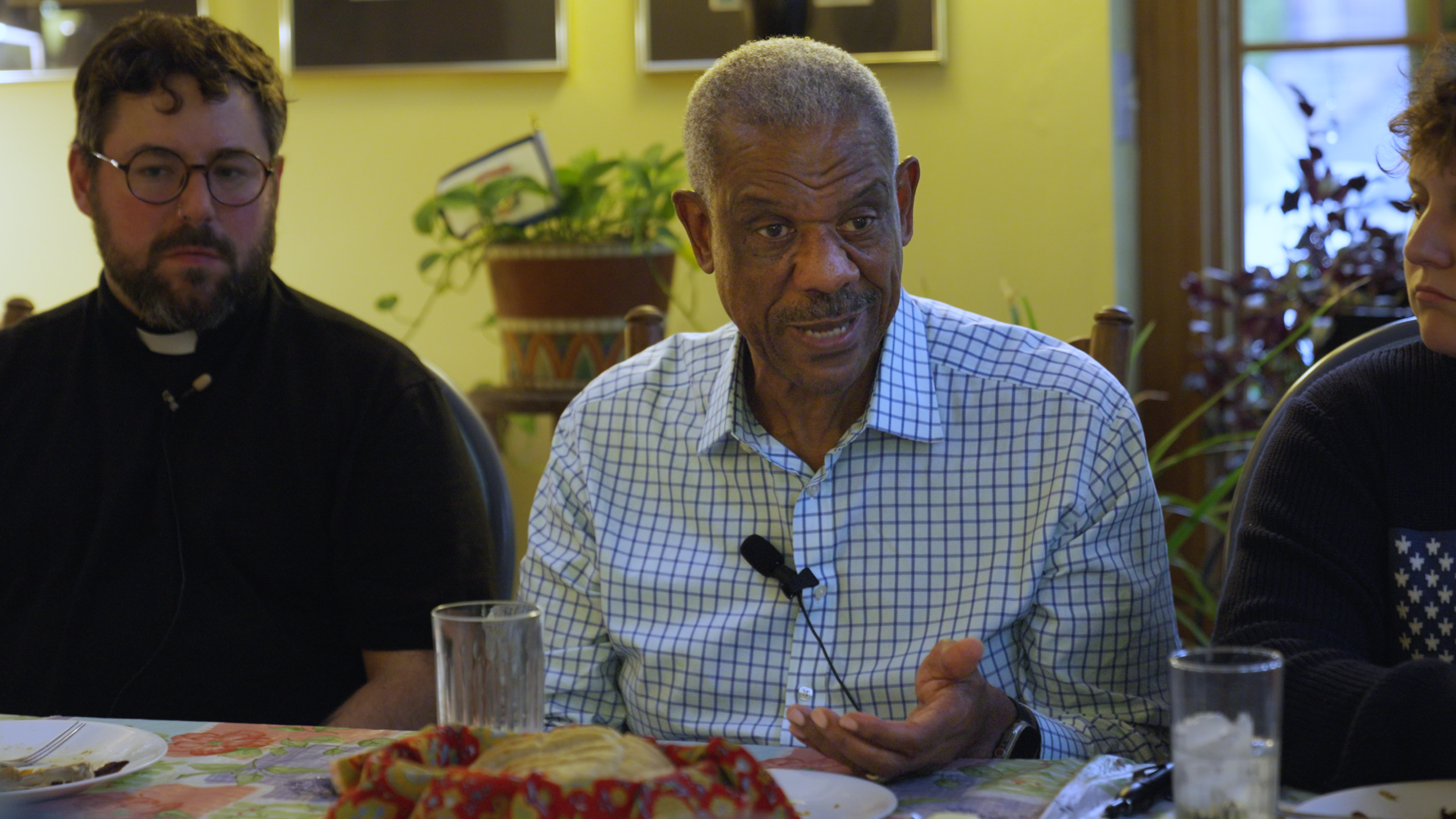 A Black man sits at a dinner table and appears to be speaking to someone off camera. He is wearing a checkered button up shirt. Next to him is a white man, a reverend, and on his other side is a person wearing a sweater.