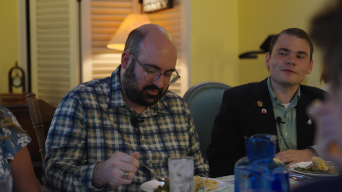 A middle-age man, balding, with brown hair looks down toward his plate at a dinner table. He wears glasses and a checkered button up shirt. Next to him is a younger man with shortly trimmed brown hair, wearing a dark blue blazer and light blue button up shirt.