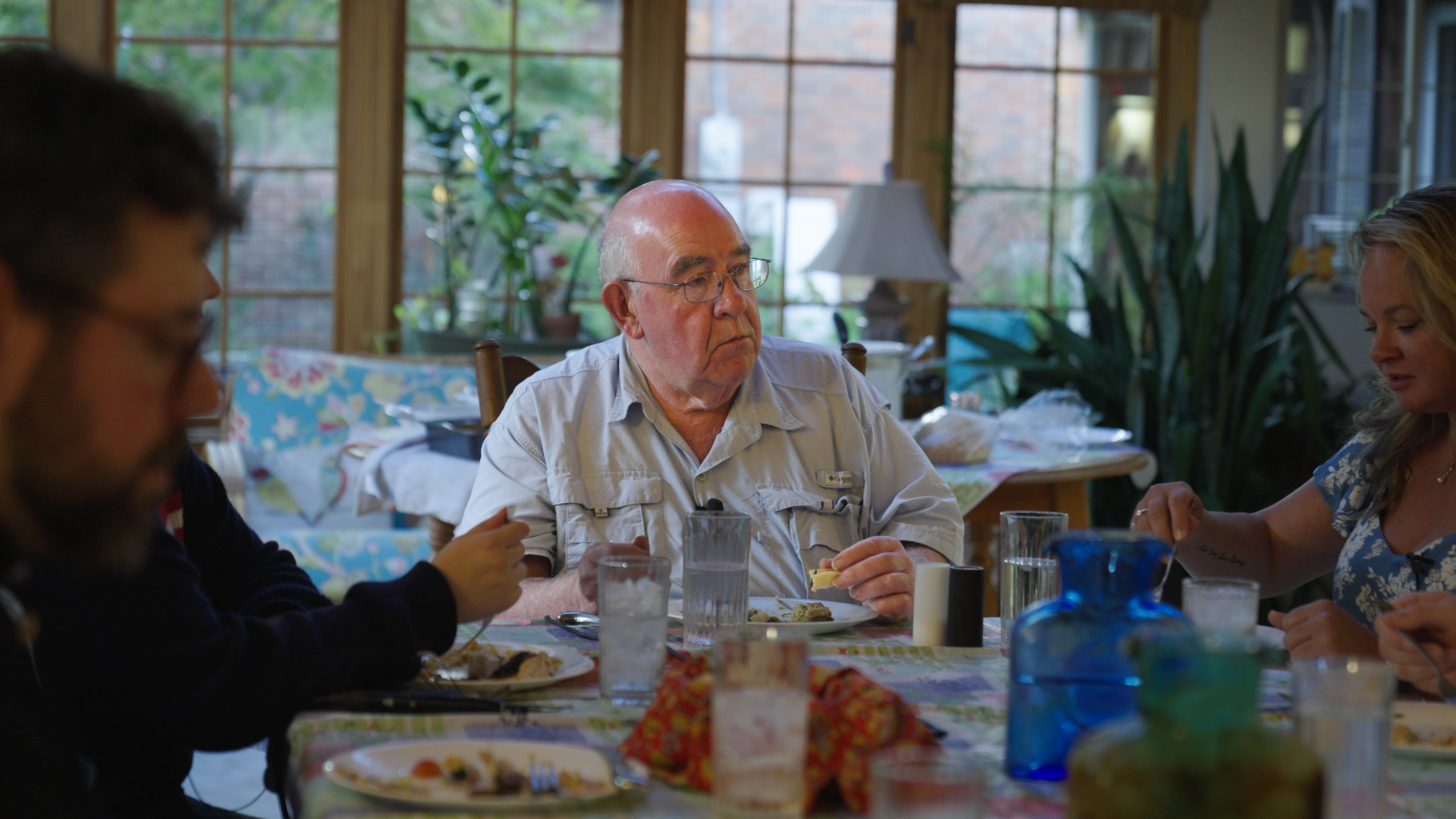 An older man sits at a dinner table and looks off camera as if listening to someone speaking. He's wearing glasses, is partly bald, and is wearing a light blue button up shirt.