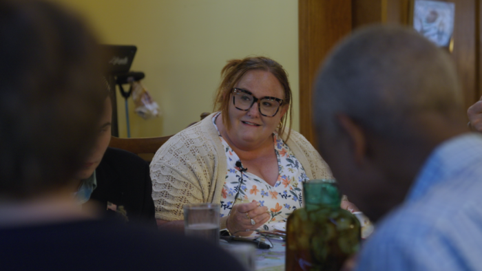 A middle-age woman with blond hair pulled back and wide-rimmed glasses sits at a dinner table. She is looking off camera and is smiling softly. She is wearing a colorful blouse with prints of flowers and a cream colored cardigan.