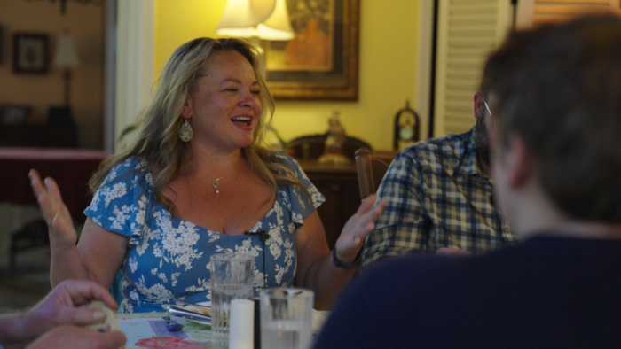 A middle age woman with blond hair and large earrings sits at a dinner table and holds her hands up as she speaks to someone off camera. She is smiling or laughing. She wears a necklace with a small pendent attached. She wears a blue and white dress.