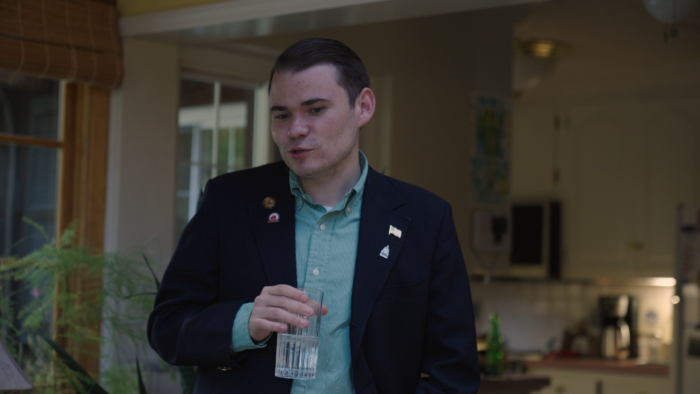 A younger man with shortly trimmed hair stands and looks down as if speaking to someone sitting down. He's wearing a dark blue blazer and light blue button up shirt. He has several pins on his lapel, including one of the American flag. In his hand is a glass of water.