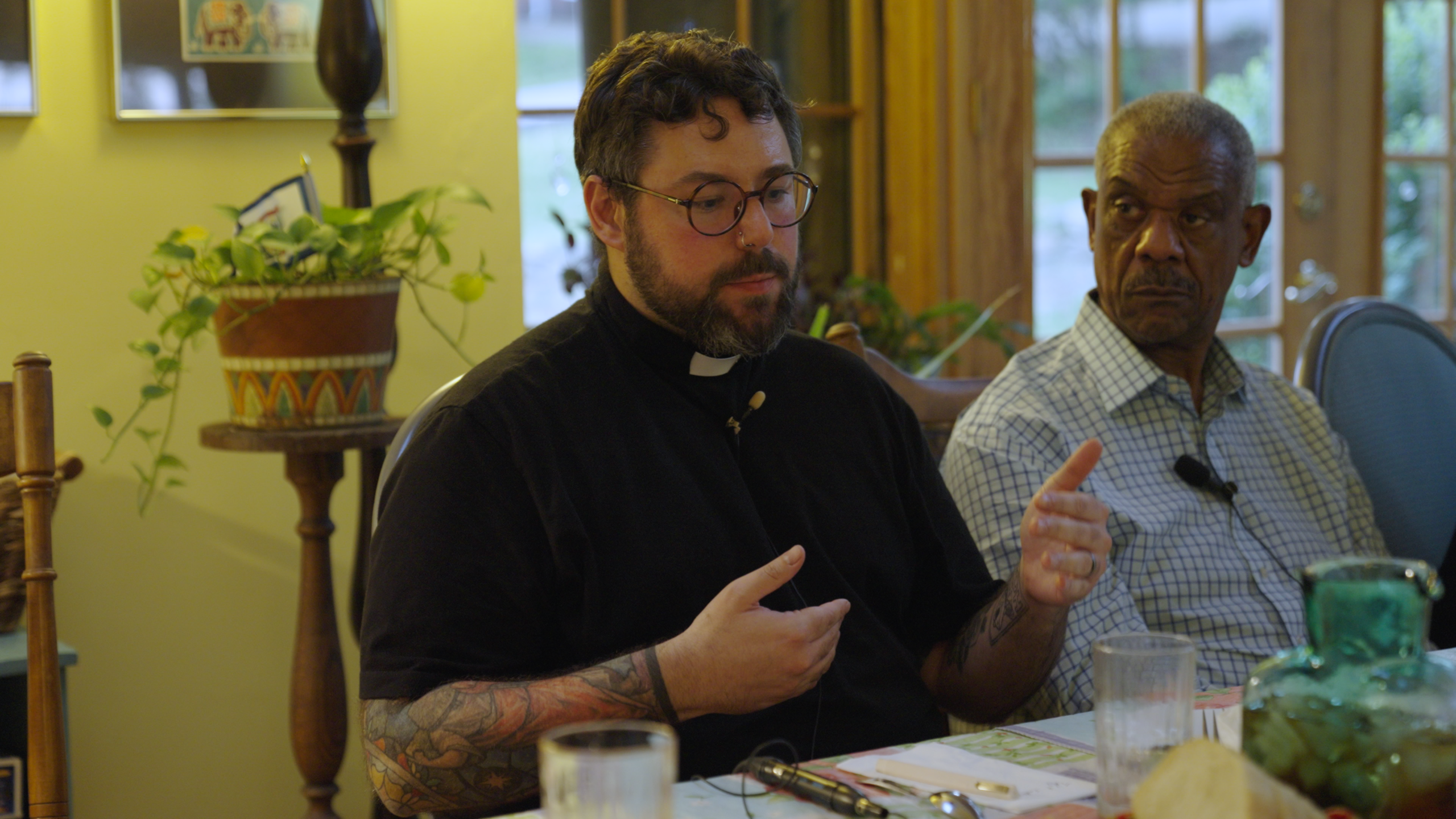A man wearing a shirt that indicates he's a reverend holds his hands up and is looking down slightly as he speaks. A man next to him looks toward him, listening. The reverend has tattoo sleeves on both arms.
