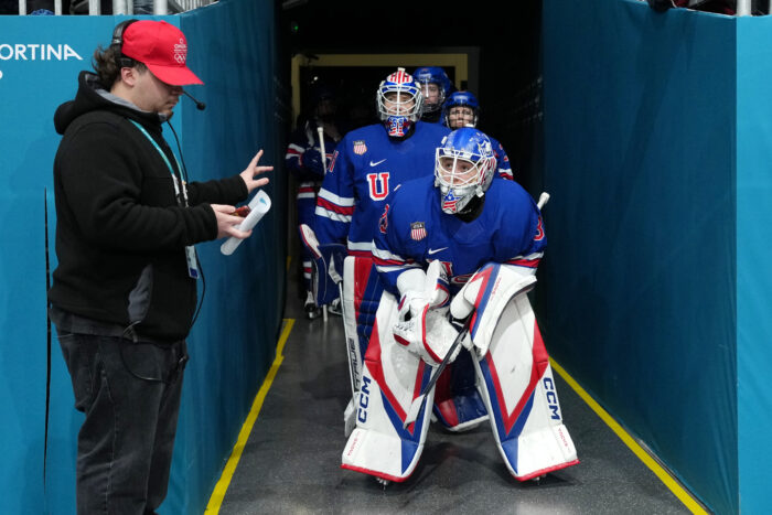 A hockey player wearing a hockey uniform that is red, blue, and white, waits to enter the ice rink for a hockey game.