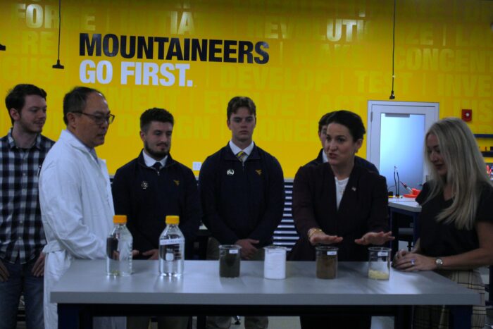 A group stands behind a table with several glass containers arrayed in front of them. One woman gestures towards containers on the far right. A figure on the left wears a white lab coat. All stand in front of a yellow wall with words painted on it, including Mountaineers Go First in blue and white.