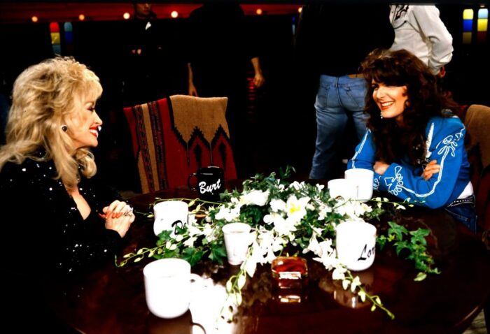 A landscape photo of two women sitting at a table across from each other. The woman on the left has blonde hair and is wearing black. The woman on the right has brown hair and is wearing blue.