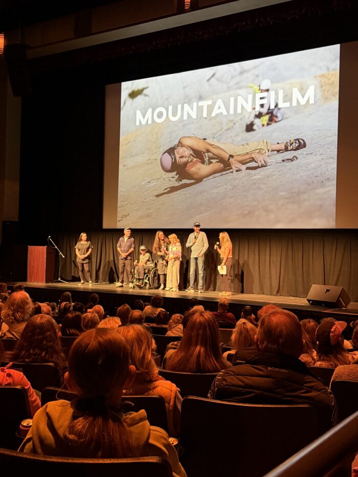 Seven people on a stage before an audience. The screen behind them shows a climber and the words Mountainfilm.