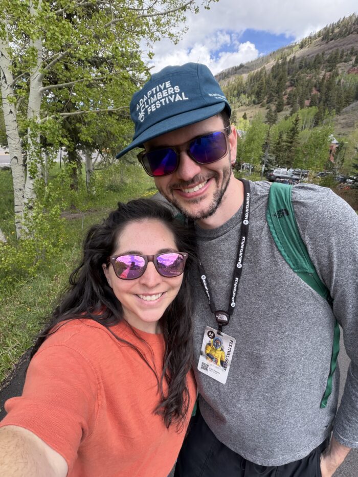 Two young people, a man and a woman, outside, dressed for hiking, take a selfie and smile for the camera.