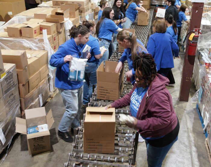 More than half a dozen young people wearing matching blue shirts are in a warehouse surrounded by pallets with cardboard boxes.
