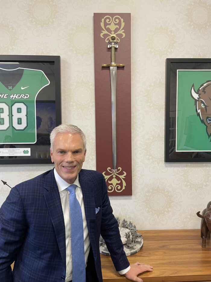 An older man with white hair stands in front of a wall featuring Marshall University memorabilia - a shirt, the Marshall bison, and a sword. The man is smiling for the camera and formally dressed in a suit and tie.