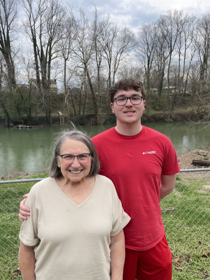 Two people, a young man and his grandmother. They are outside against a backdrop of grass and a river or pond. The boy is wearing a red shirt, and the woman is in a tan shirt. They are both wearing glasses, have brown hair, and are smiling at the camera.