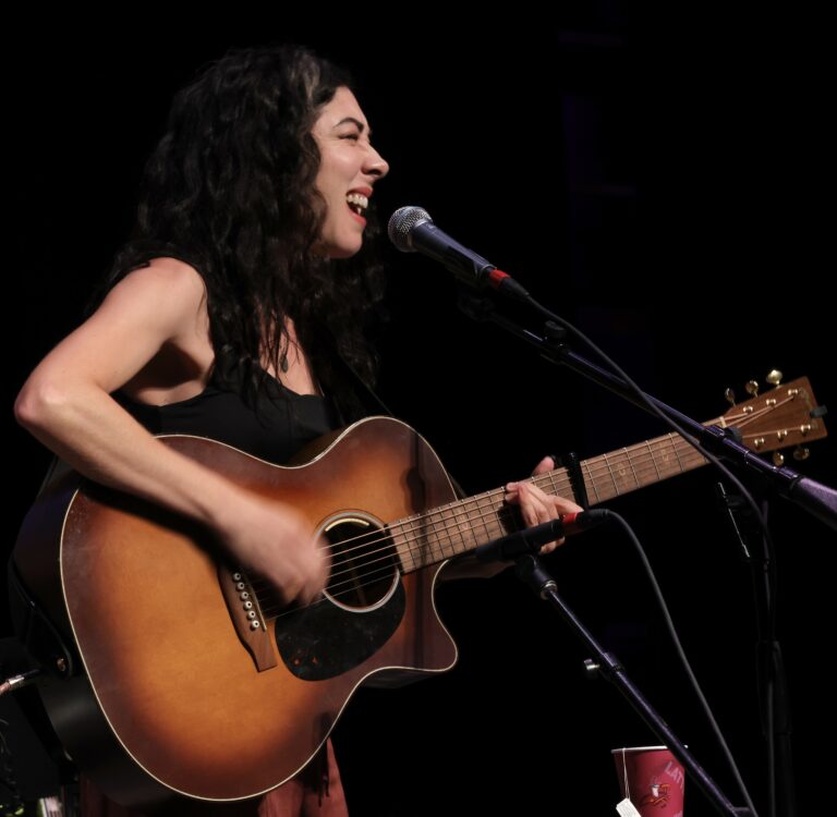 a woman with long dark hair plays the guitar and sings into a mic