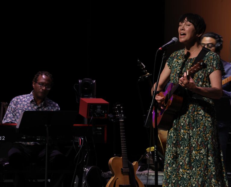 woman sings and plays the guitar and man in background plays the piano