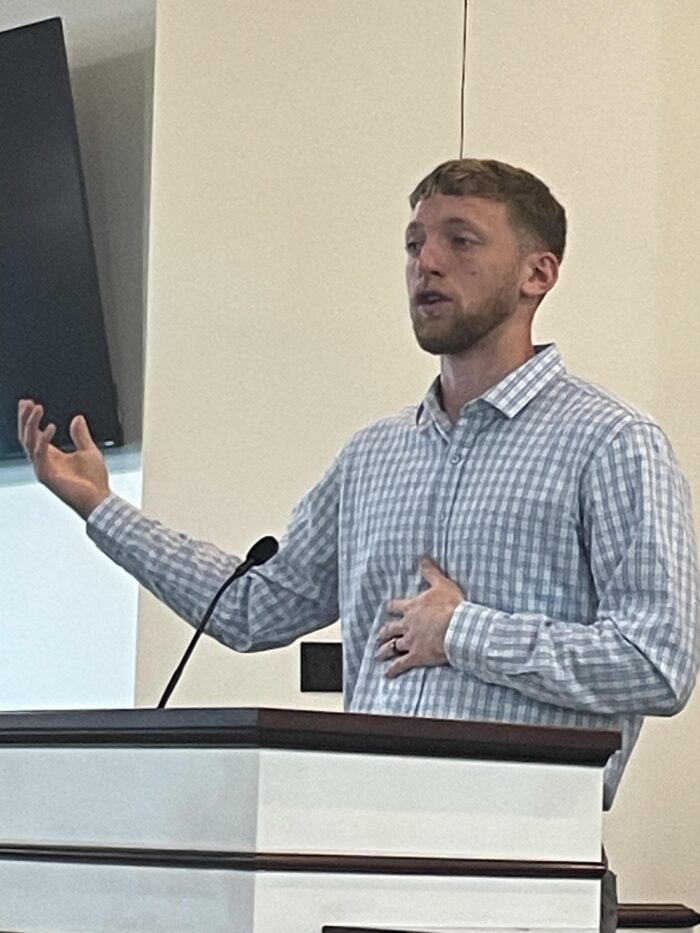 A young man with brown hair and beard, wearing a button up shirt, speaks at a podium.