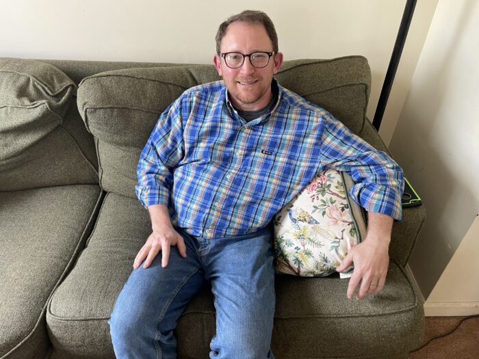 A middle age man with brown hair and beard, wearing glasses and a blue plaid button shirt, looks up toward the camera smiling. He is sitting on a couch with is arm hanging over a pillow.