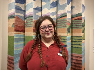 A landscape portrait of a young white woman standing in front of a tapestry. She is wearing a red sweater and braids.