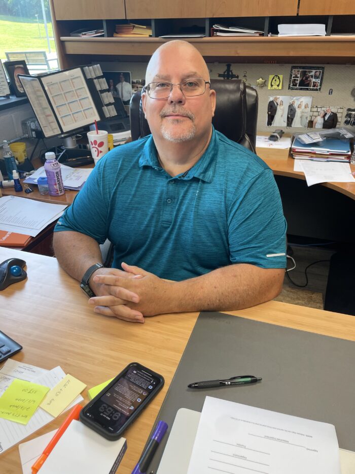 An middle age man sits at his desk and looks up toward the camera, smiling softly. He is wearing glasses and a blue polo shirt.