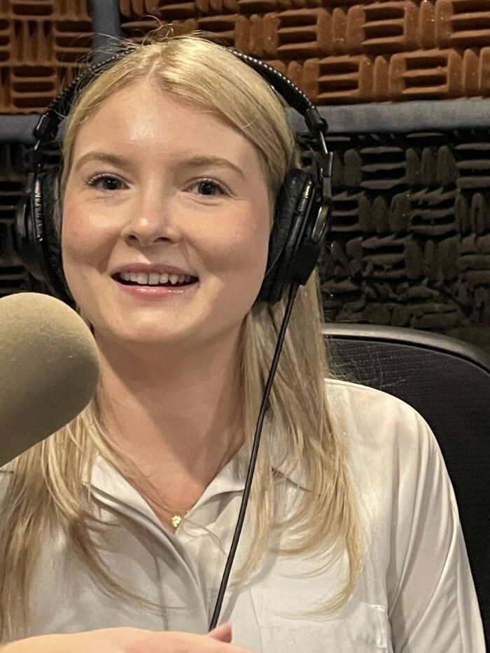 A young woman, blond and in a white dress shirt, sits in a recording studio. She has headphones on and speaks into a microphone while looking toward the camera.