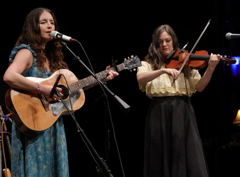 a woman plays a guitar next to a woman playing the fiddle
