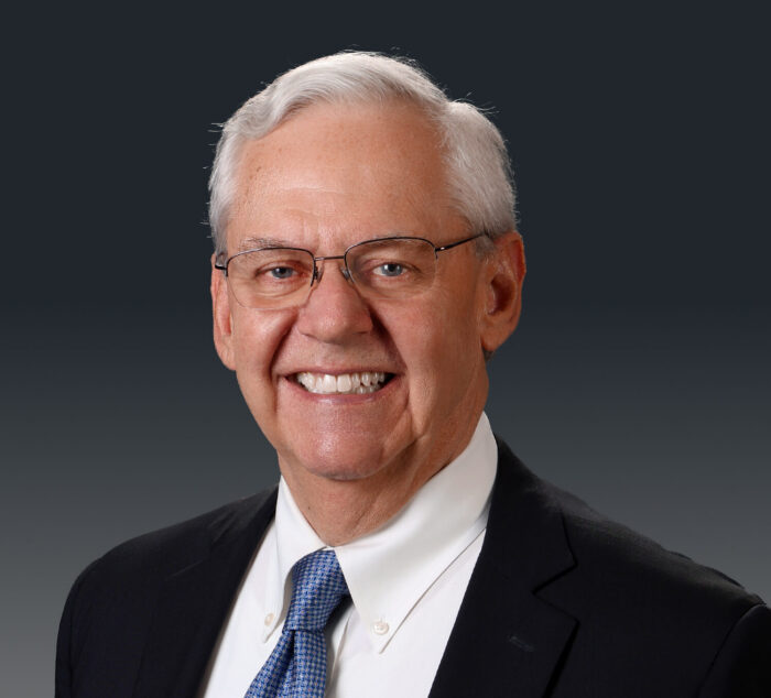 A professional headshot of an older man with white hair formally dressed in a suit and tie. He smiles for the camera and is wearing glasses.