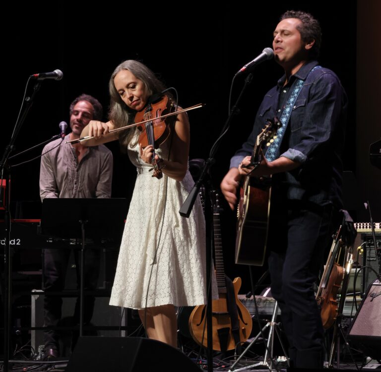 a man plays the guitar and a woman with grey hair in a white dress plays the fiddle