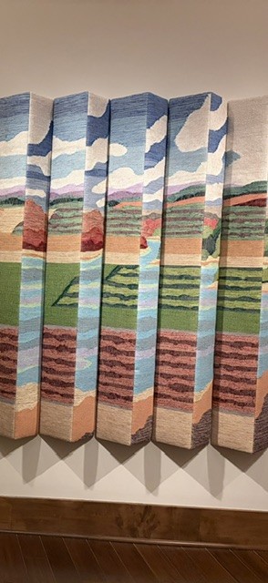 A vertical photo of a tapestry. The artwork illustrates farm land.