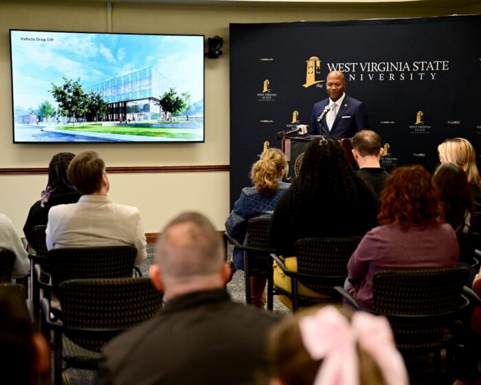 A seated crowd in a room faces a flatscreen mounted on a wall displaying an artist's render of a glass and steel building. A man stands at a lectern to the right of the television in front of a dark blue backdrop.