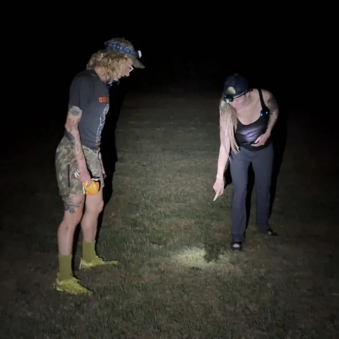 Two women stand outside at night wearing headlamps. They appear to be tracking something.