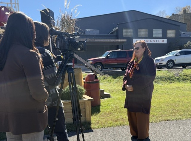 A woman in a brown suit stands before several television cameras and microphones. Behind her is a grassy field and a gymnasium.