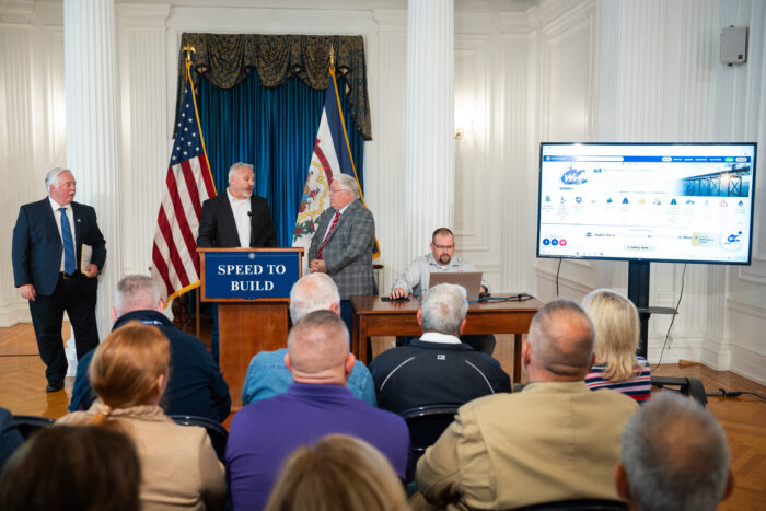Three men in suits stand behind a lectern with a sign reading "Speed of Business" in a large white room next to a man sitting at a wooden table in front of a laptop with a website displayed on a flatscreen.