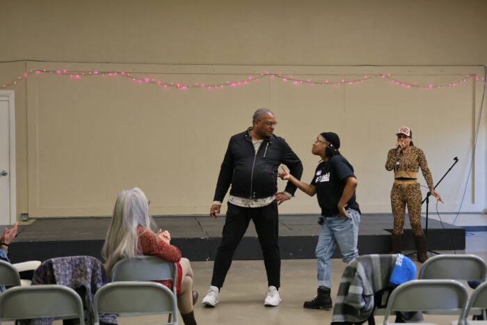 A photo of three people standing in front of a stage. Two of them are looking at and talking to each other, while another person talks into a microphone in the right corner. There is one person sitting left of the performers with their back facing the camera.