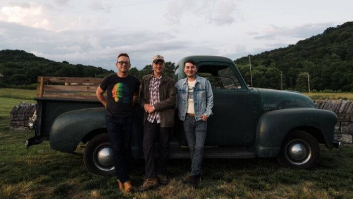 Three white men standing in front of a truck. They are surrounded by mountain foothills and greenery.