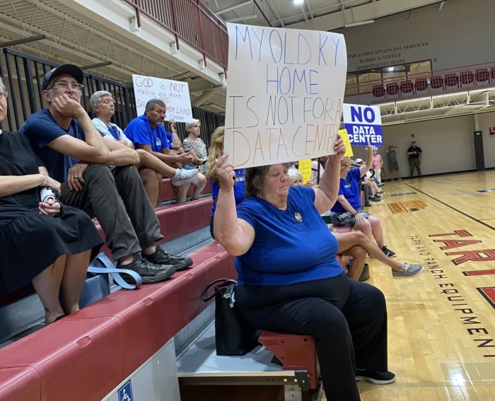 People sitting on red benches inside a gymnasium.  They are holding signs and wearing blue shirt. The woman in the foreground is holding a sign that reads, "My old KY home is not for a data center."