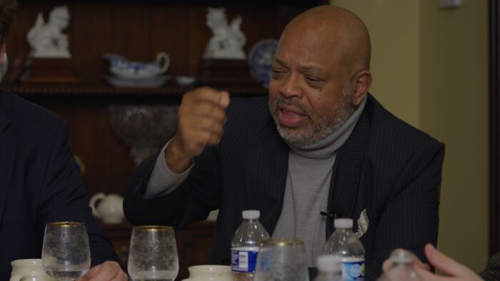 A Black man speaking while sitting around a dinner table.