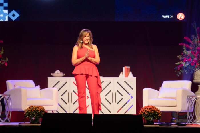 A woman in a sleeveless red jumpsuit stands alone on stage with an interview set including several chairs behind her.