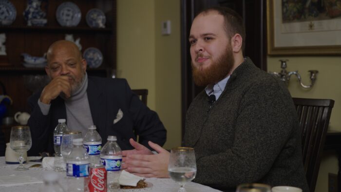 Two men sit at a dinner table. One Black and the other white.