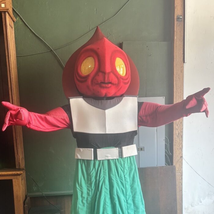 A handmade stand-up of the Flatwoods Monster. It has a pointy red head with big yellow eyes, outstretched red arms, and a white and green outfit.