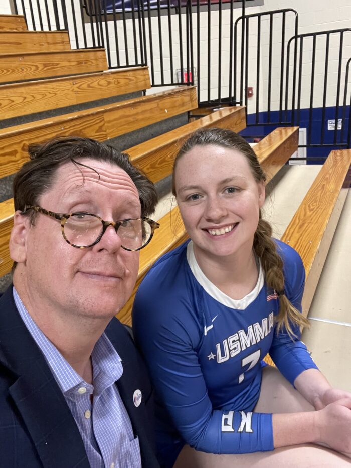Two people. A man and a young woman. Both have brown hair. The man is wearing glasses. They smile and took towards the camera for a selfie. They're sitting on bleachers in a gymnasium.