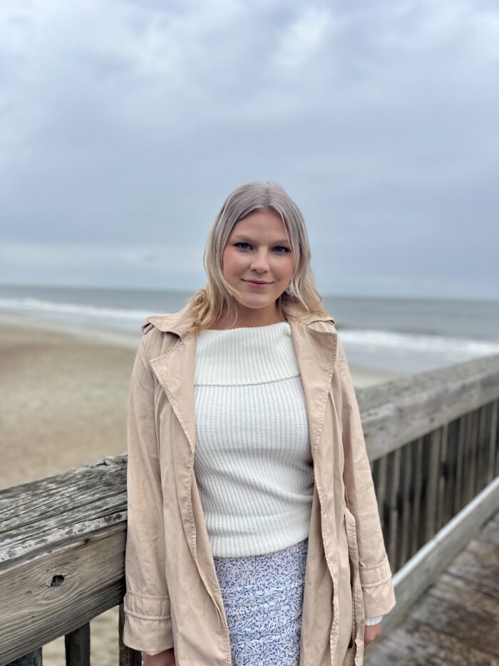 A young woman with blond hair stands on a beach against an overcast sky. She is wearing a tan jacket and white sweater.