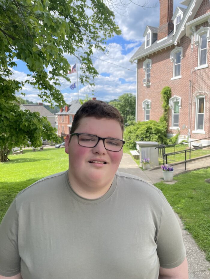 A young man smiles for the camera. He has short, brown hair and is wearing glasses. He's standing outside on a clear, sunny day. Behind him is a brick building.