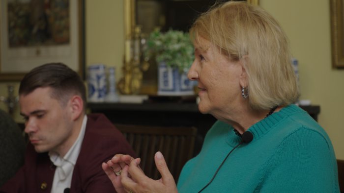 An older woman with blond hair sitting at a dinner party, speaking. Next to her is a young man with brown hair.